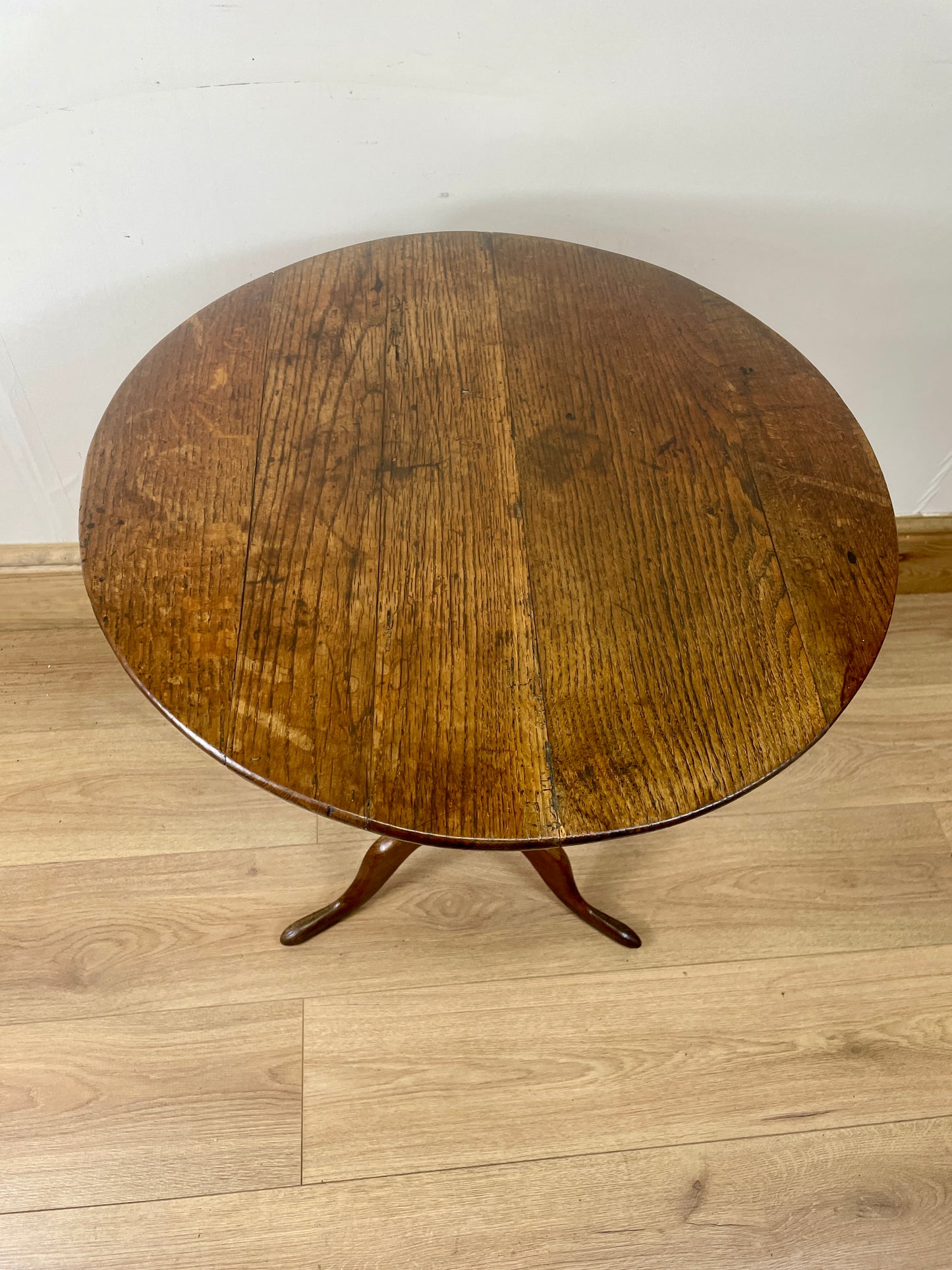 Round wooden table with a single leg on a light wood floor against a white wall.