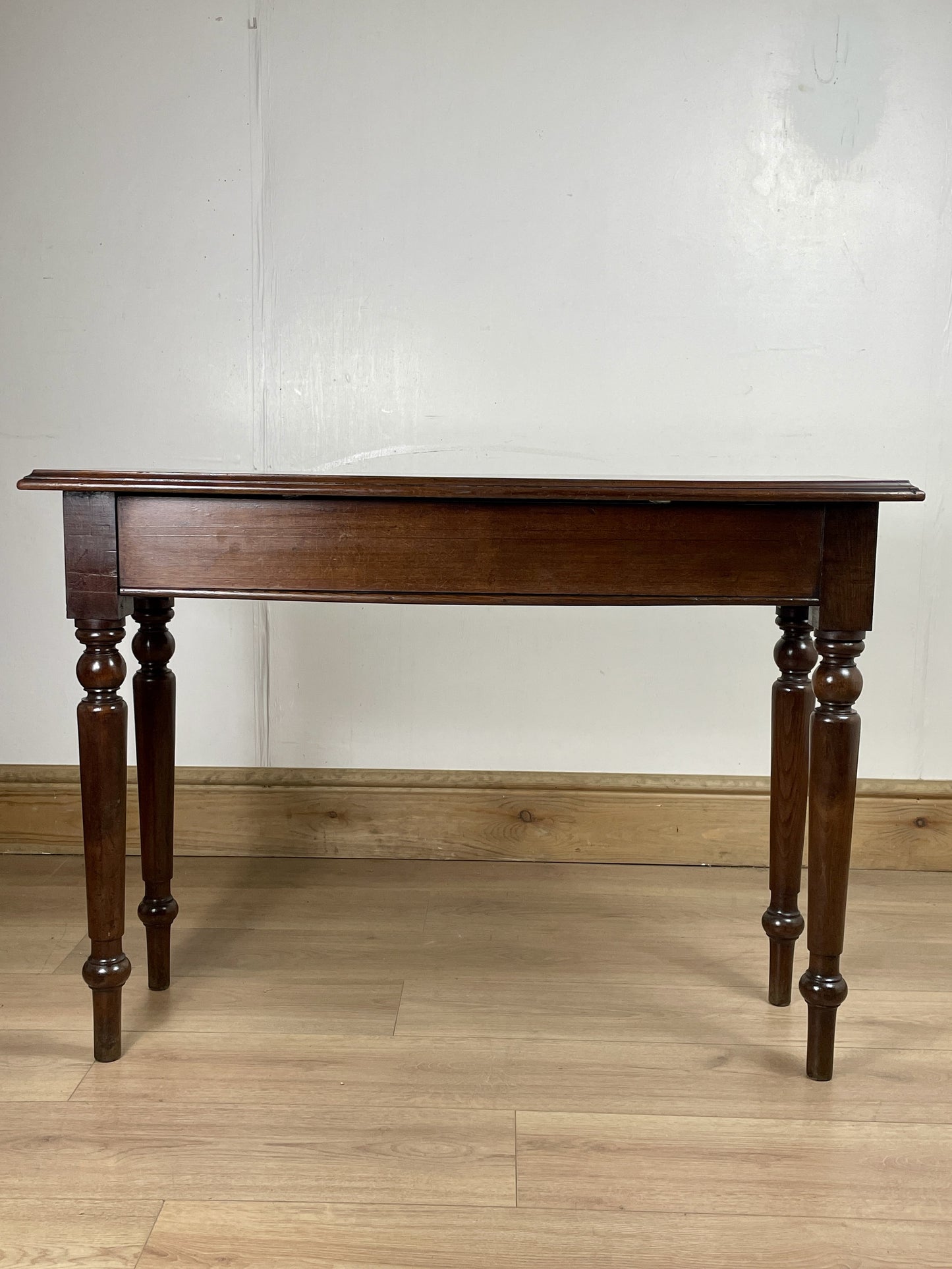 Wooden table with a single drawer on a wooden floor against a white wall.