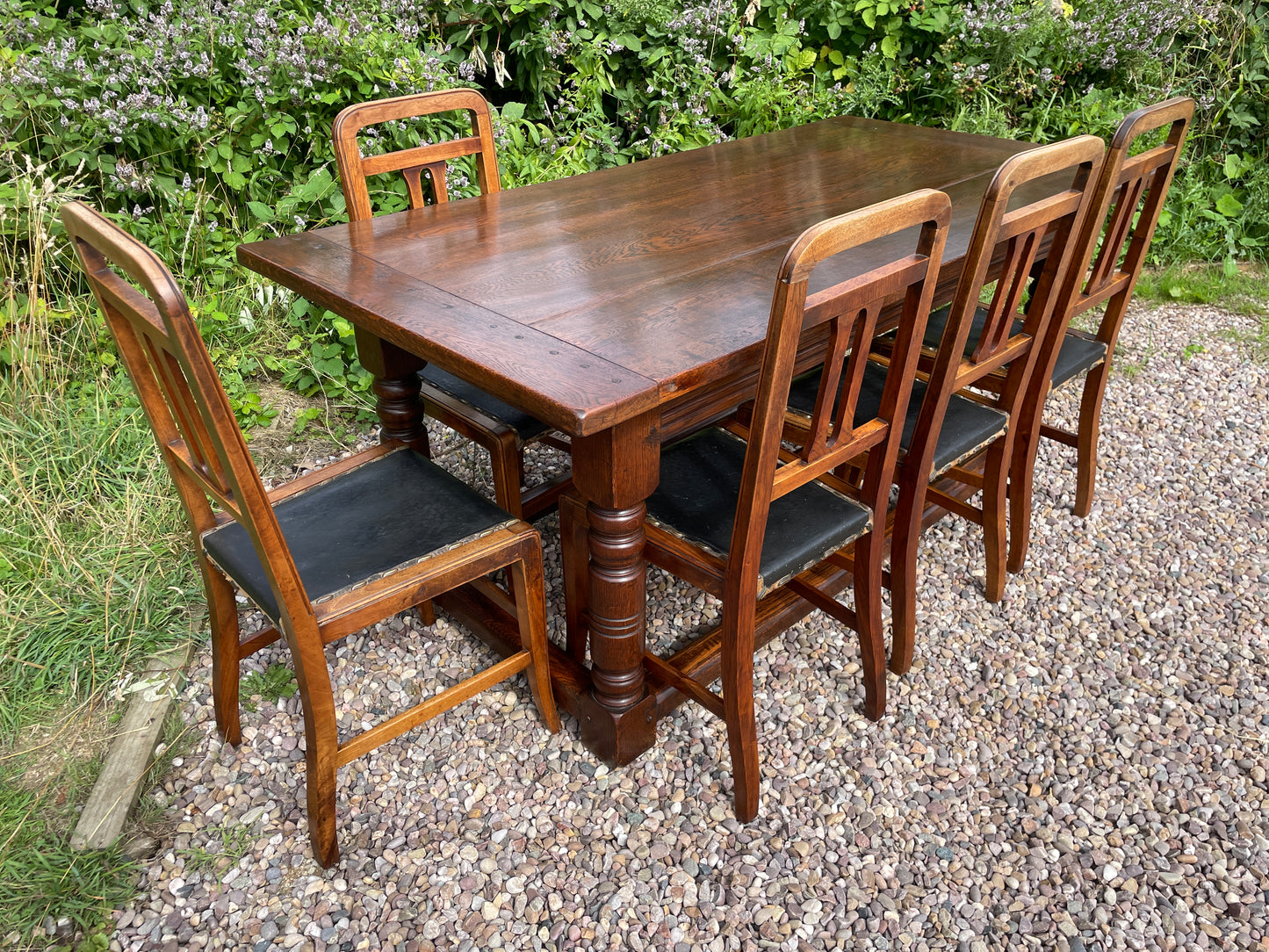 Wooden dining table with six chairs on a gravel surface with greenery in the background