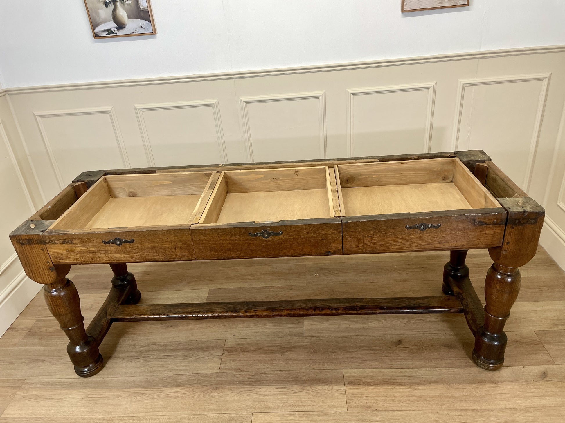Wooden table with three drawers on a wooden floor against a white paneled wall.
