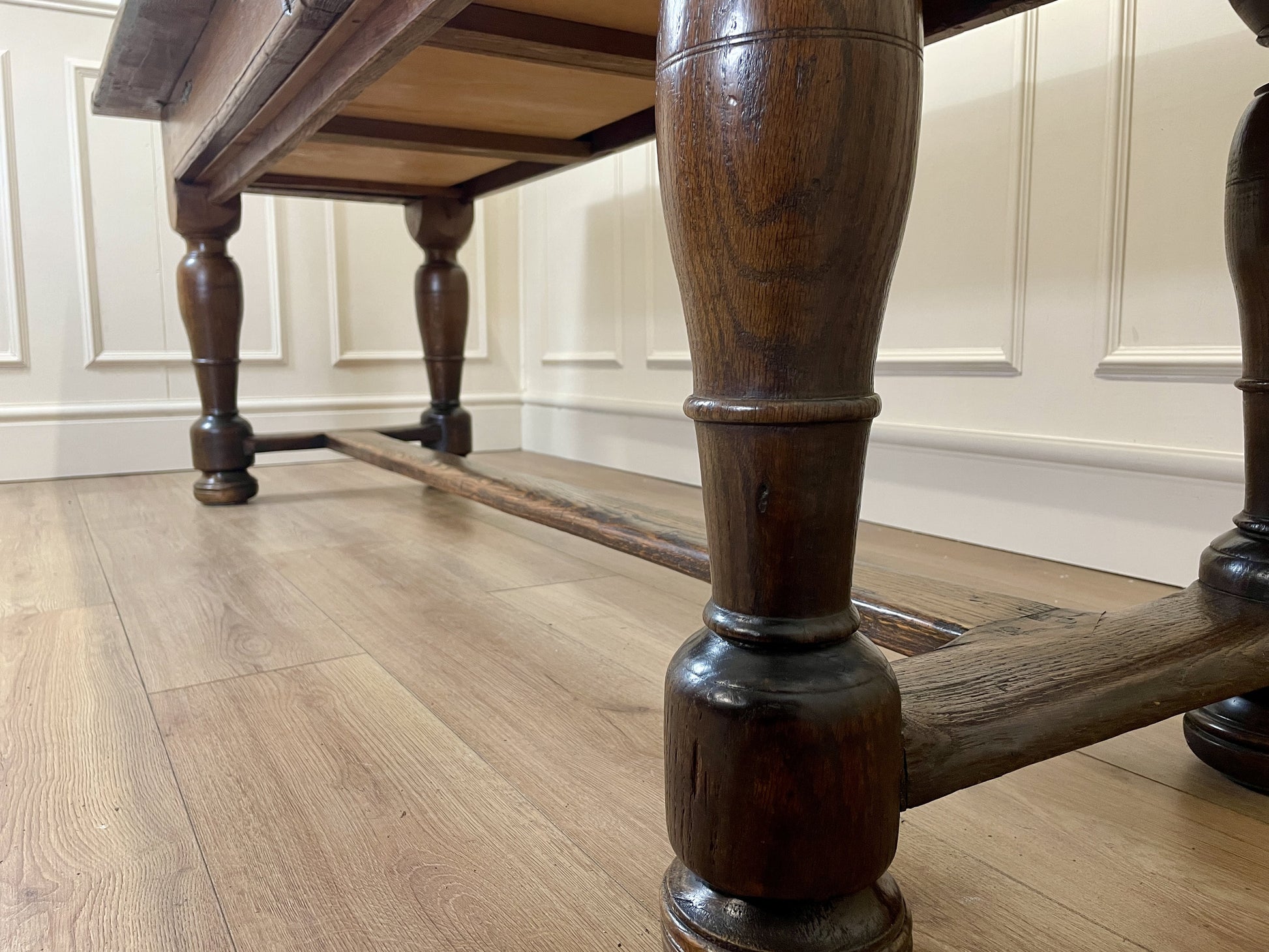 Close-up of a wooden table leg on a wooden floor with white paneled walls in the background.