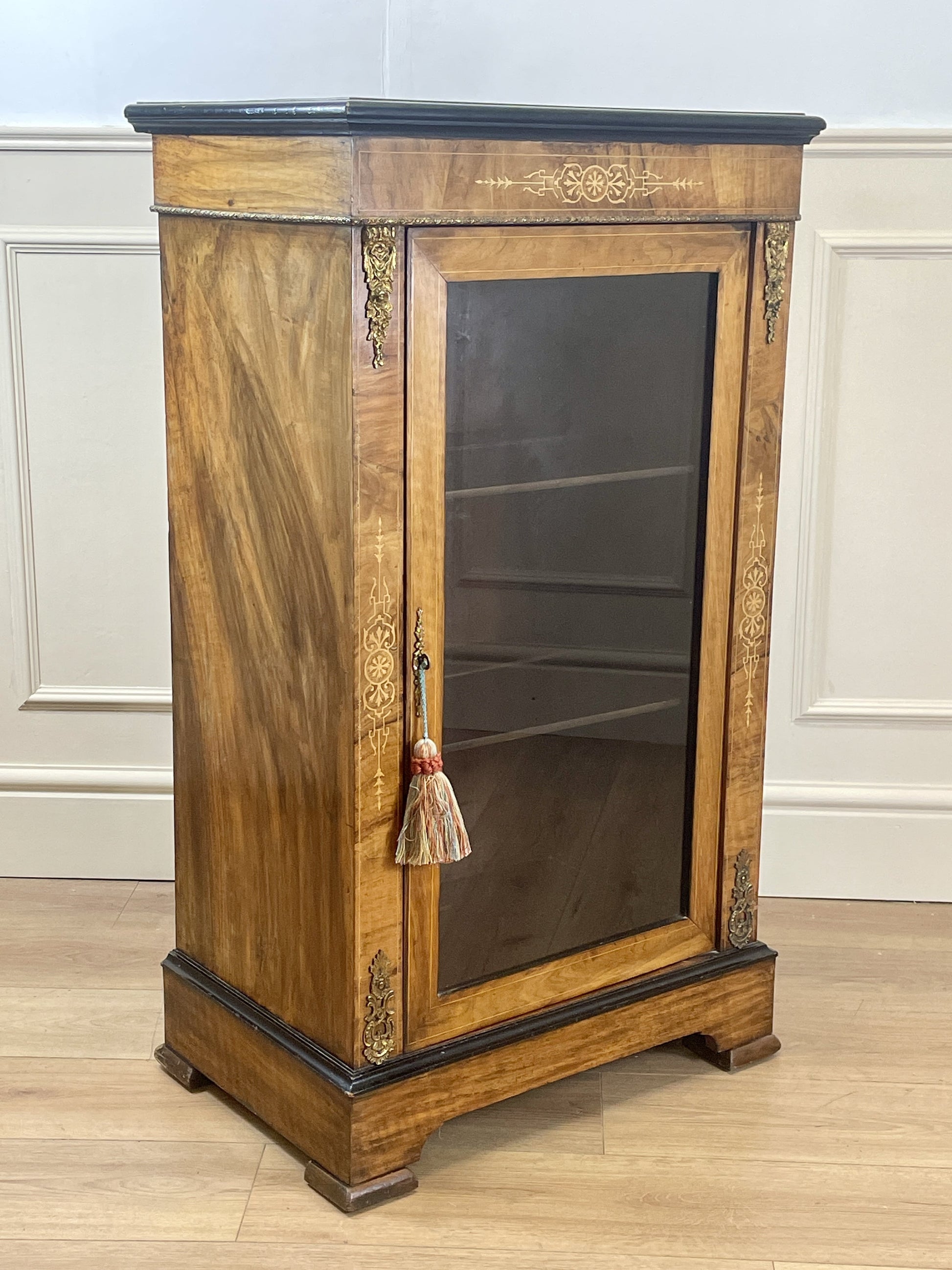 Wooden display cabinet with glass door on a wooden floor against a white paneled wall.