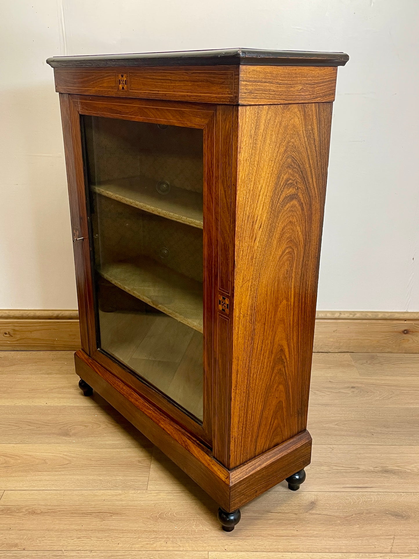 Wooden bookshelf with glass door on a wooden floor against a white wall