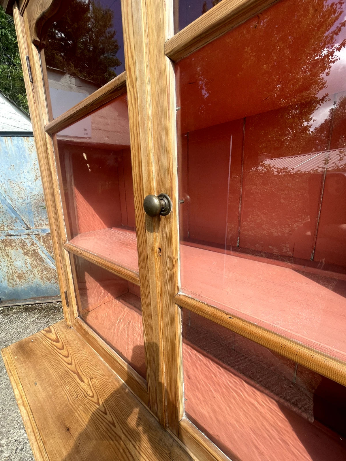 Wooden display case with glass panels and a metal handle, reflecting trees and a rusted surface.
