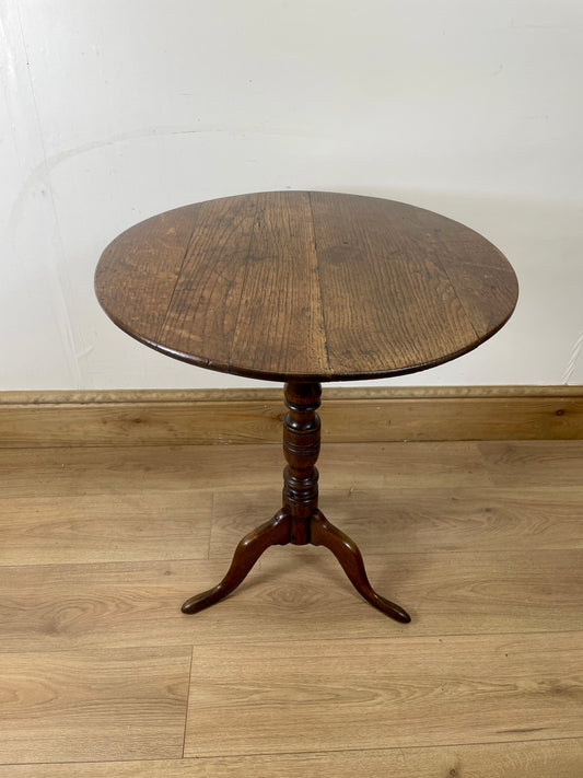 Round wooden table with a pedestal base on a wooden floor against a white wall.