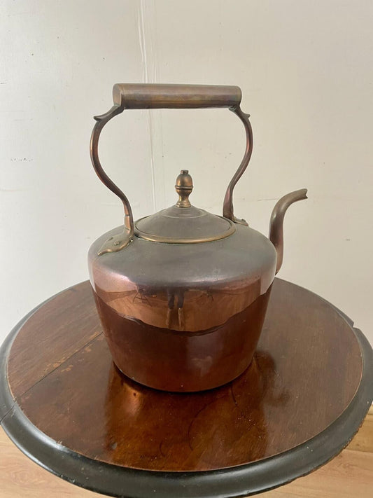 An antique copper kettle with a rounded body and a long spout, featuring a curved handle and a decorative finial on the lid, displayed on a round wooden table.