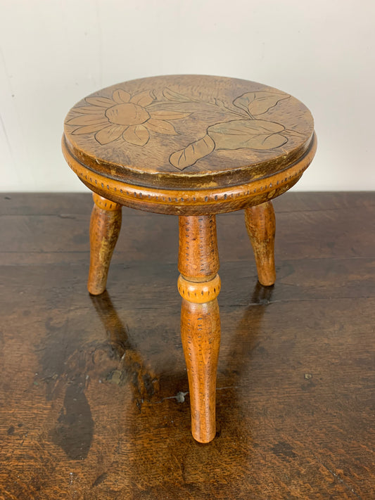 Antique wooden stool with floral carvings on the seat and three legs, placed on a dark surface.