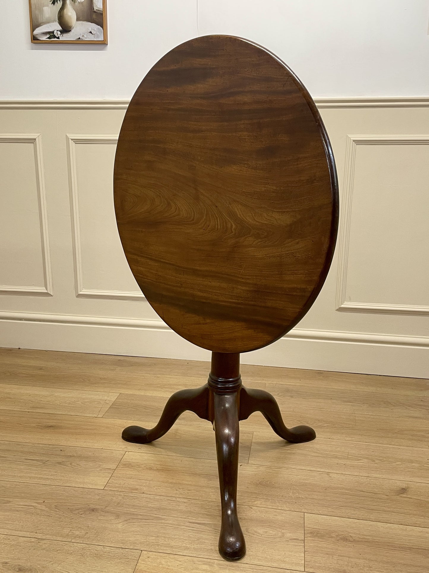 Wooden round table with a pedestal base on a wooden floor against a white paneled wall.