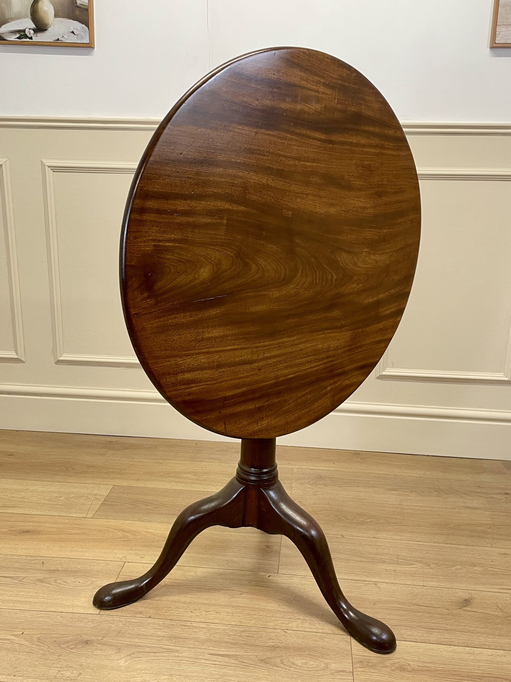 Wooden round table with a pedestal base on a wooden floor against a white paneled wall.