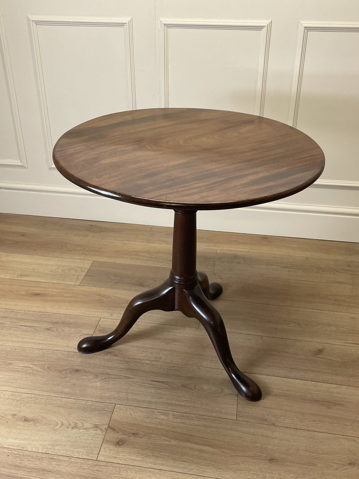 Round wooden table with a pedestal base on a wooden floor against a white paneled wall.