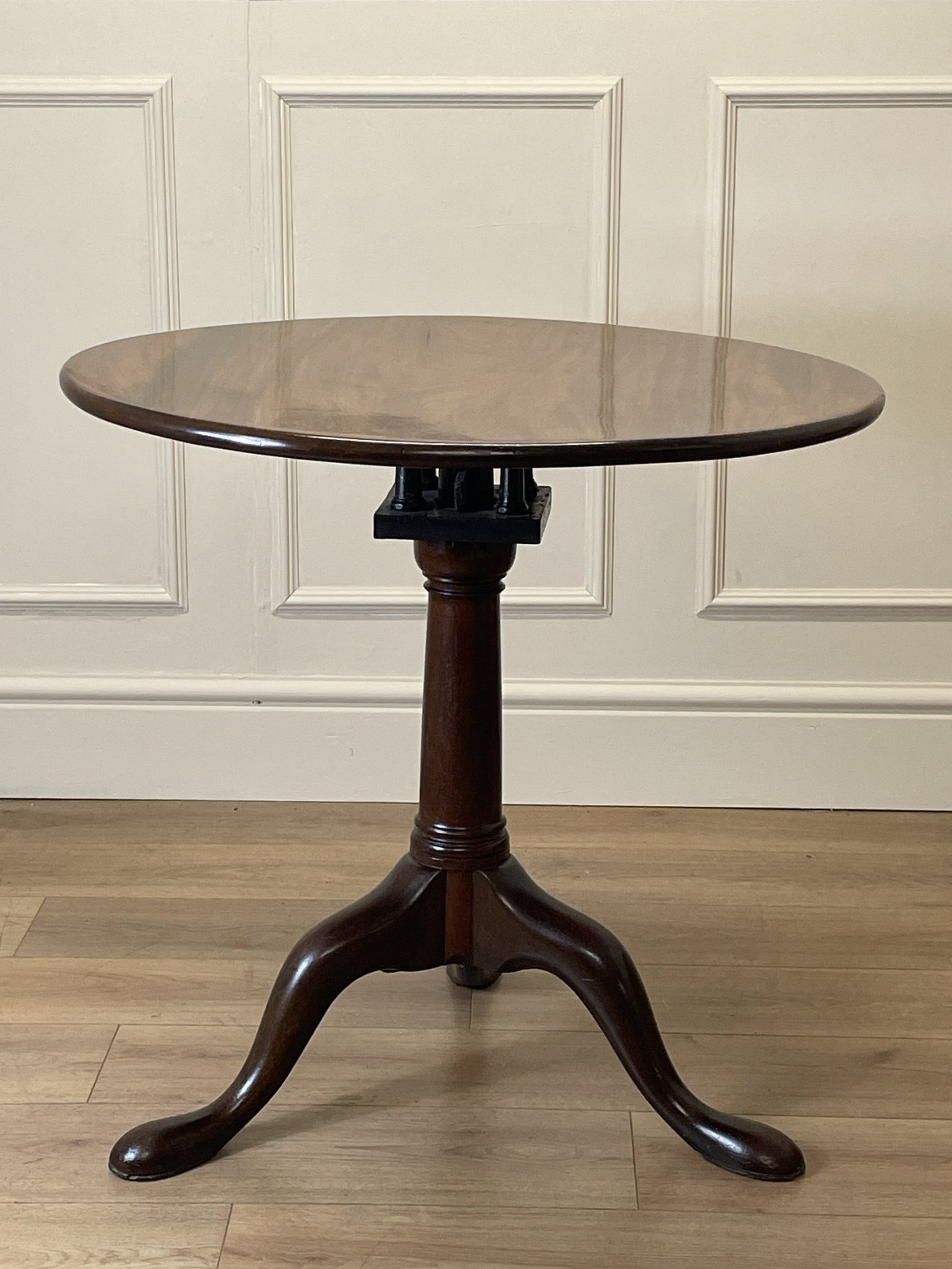 Round wooden table with a pedestal base on a wooden floor against a white paneled wall.