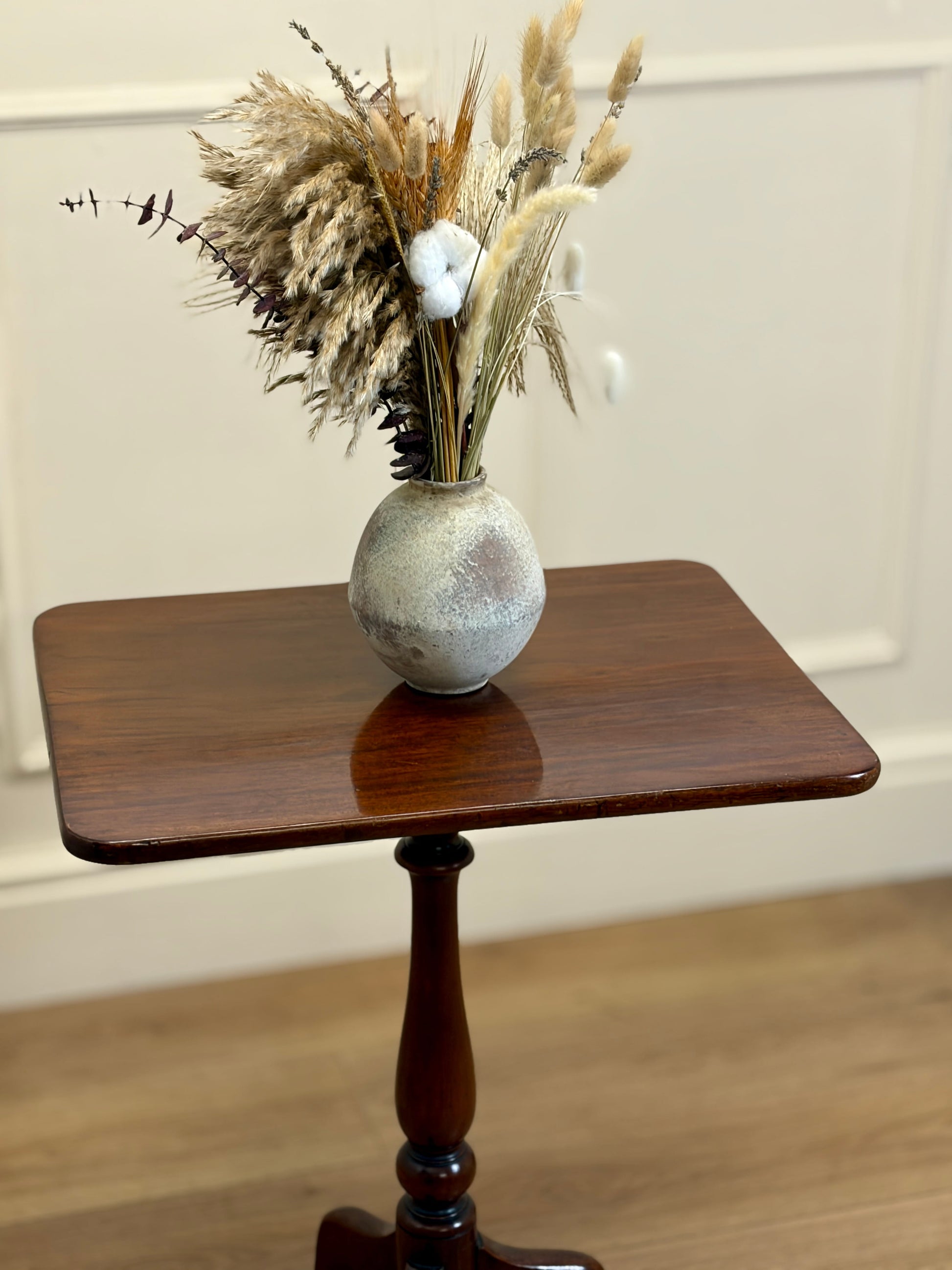 Small vase with dried plants on a wooden side table against a white wall.