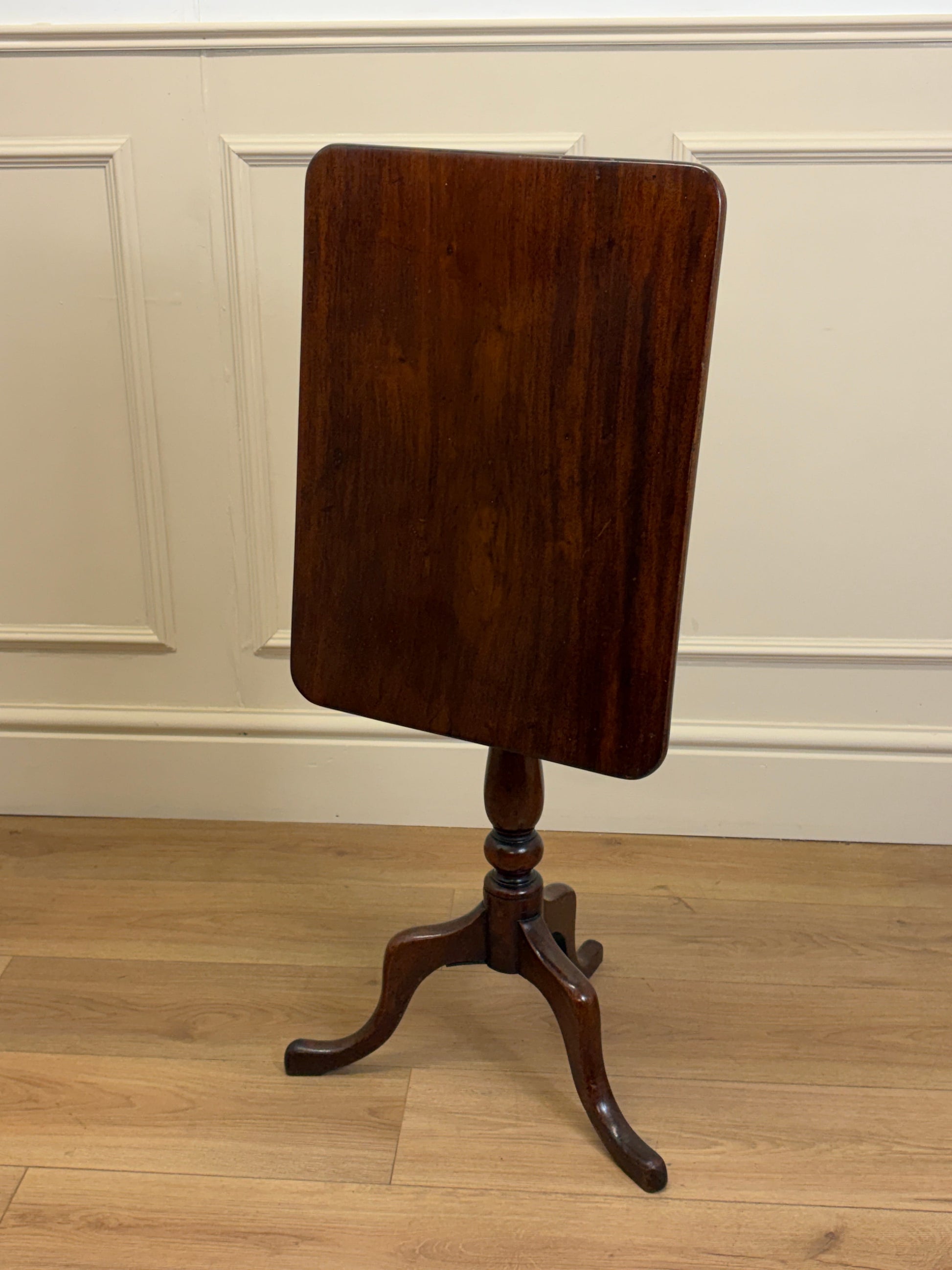 Wooden table with a single leg on a wooden floor against a white paneled wall.