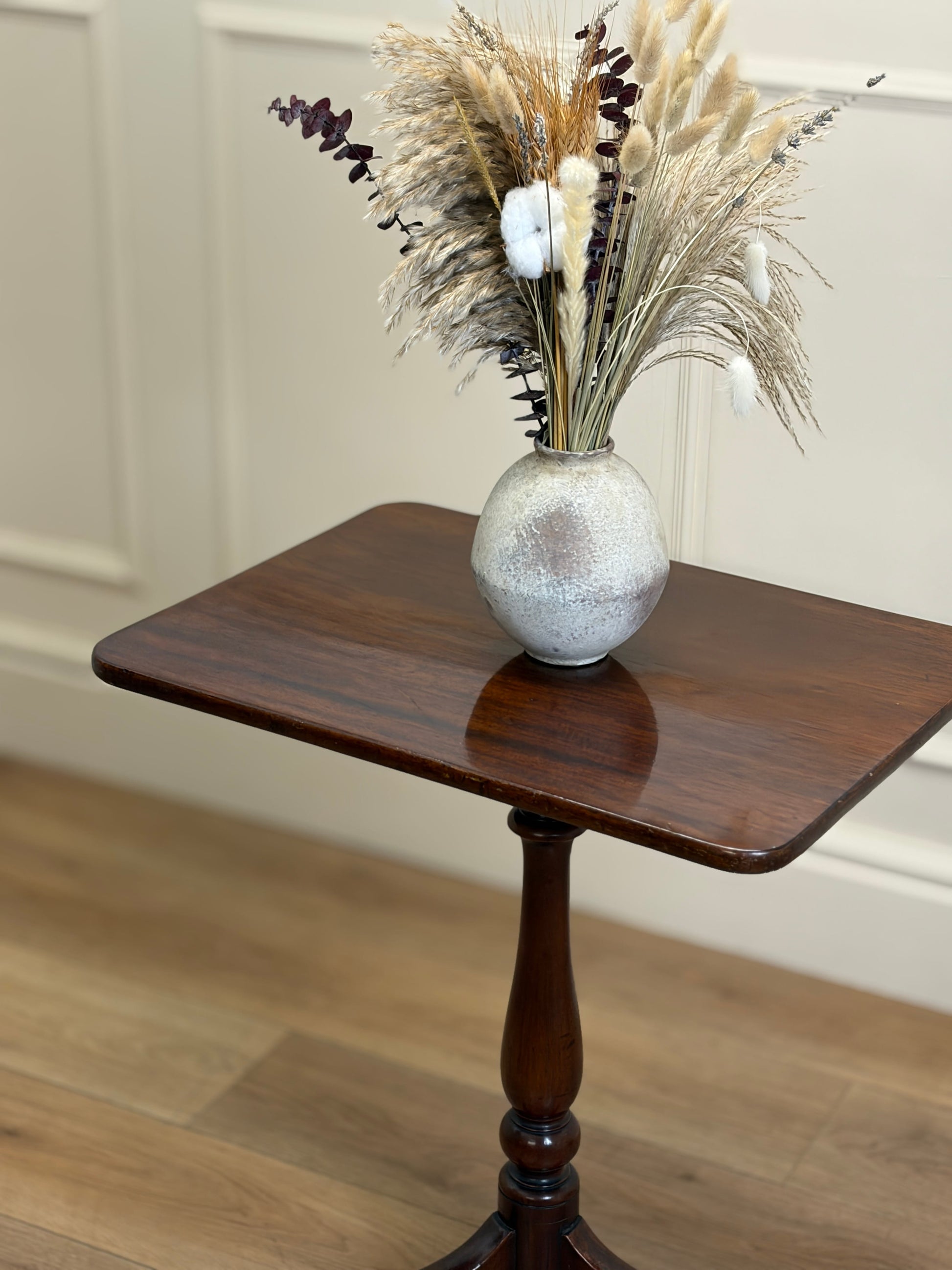 Decorative vase with dried plants on a wooden side table against a neutral wall.