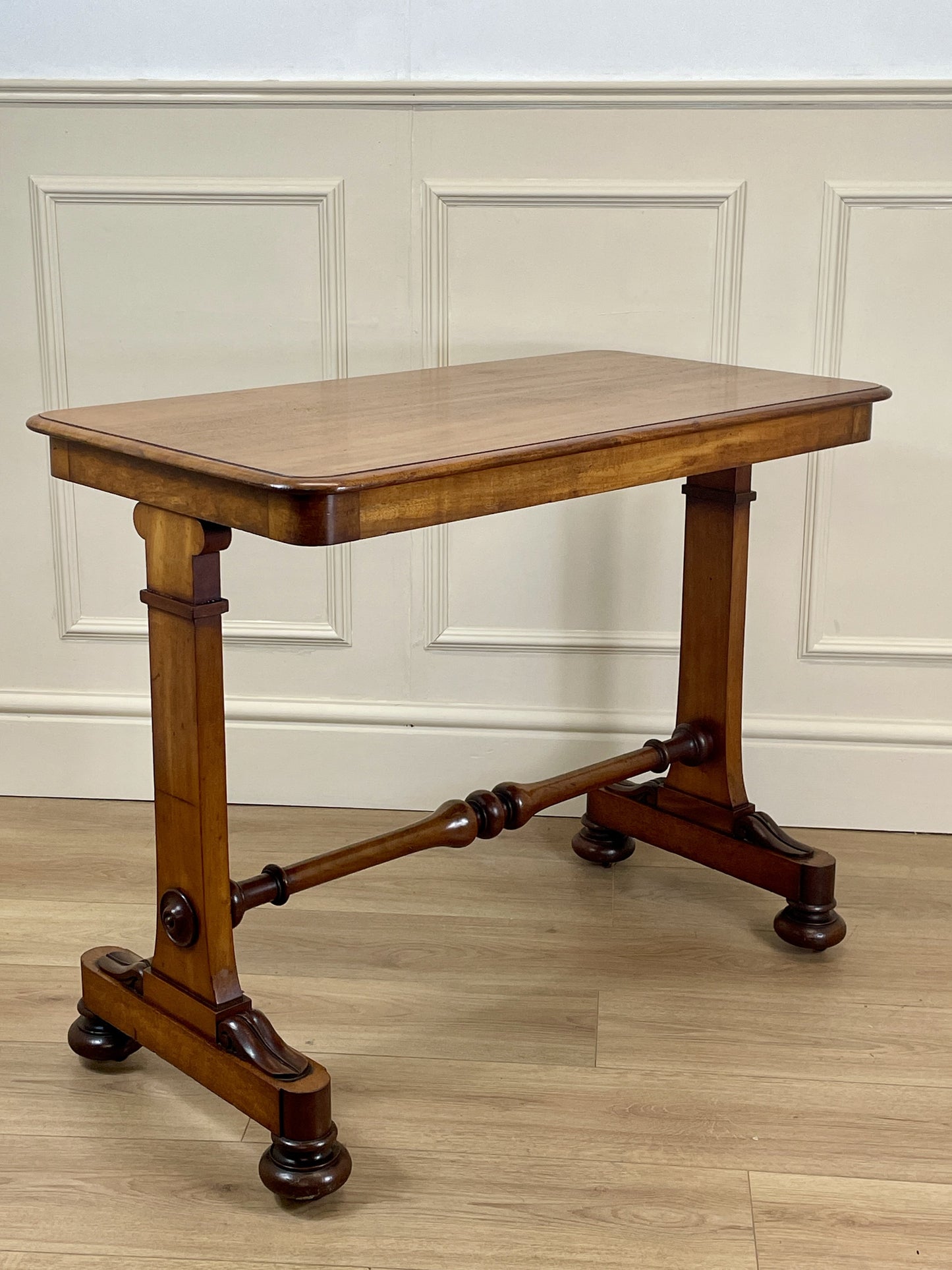 Wooden table with a classical design on a wooden floor against a white paneled wall.