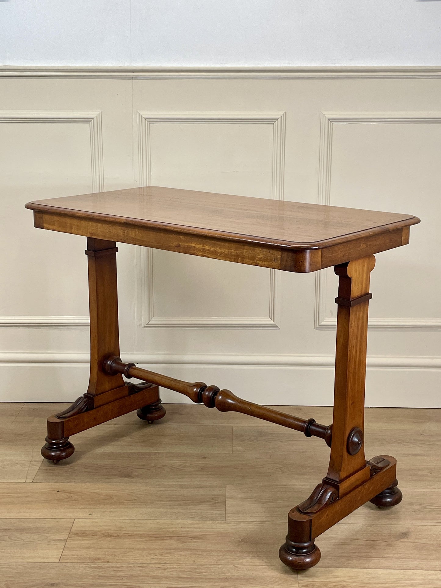 Wooden table with a classical design on a wooden floor against a white paneled wall.