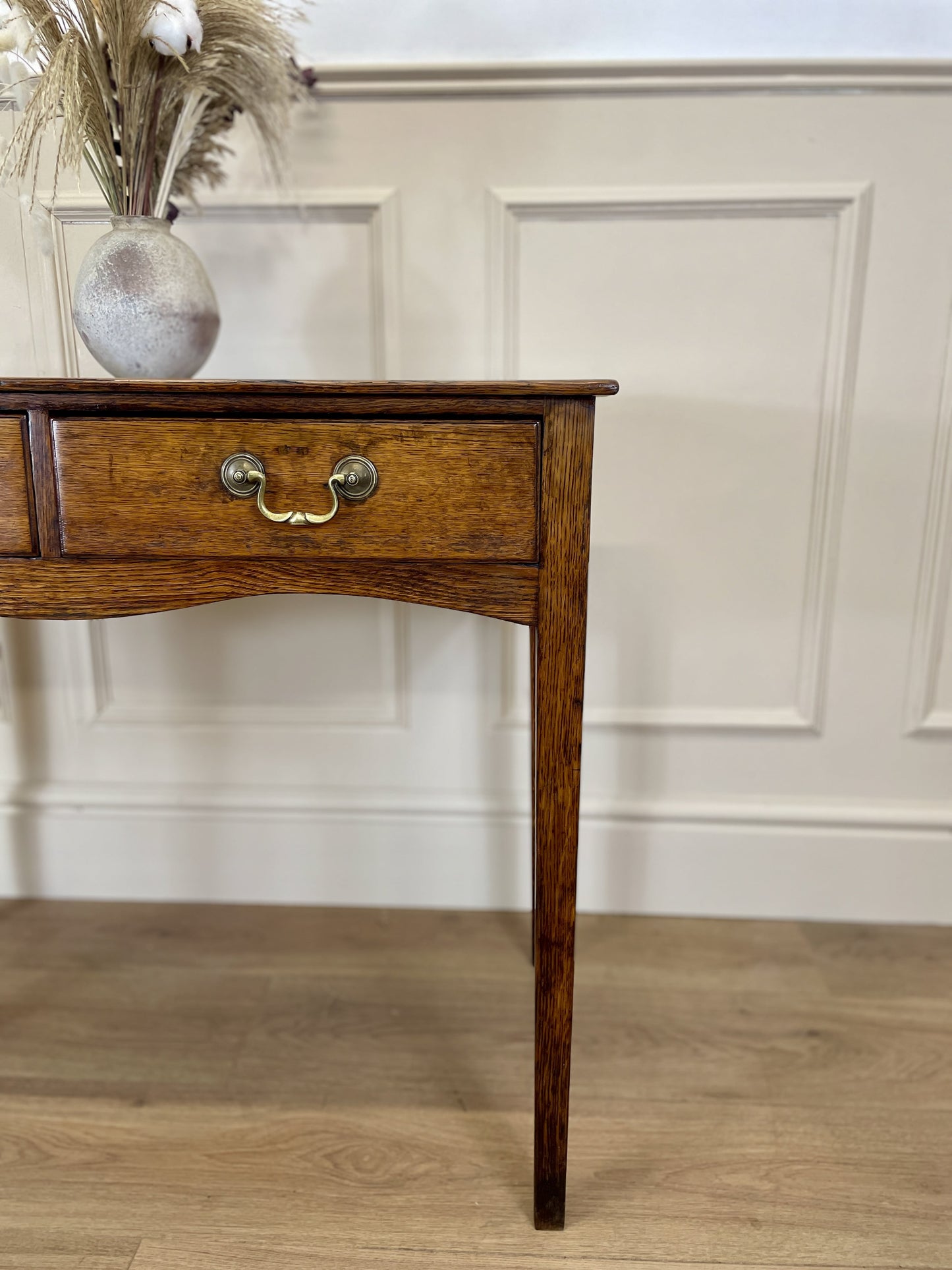 Wooden side table with a drawer and decorative vase against a white paneled wall.