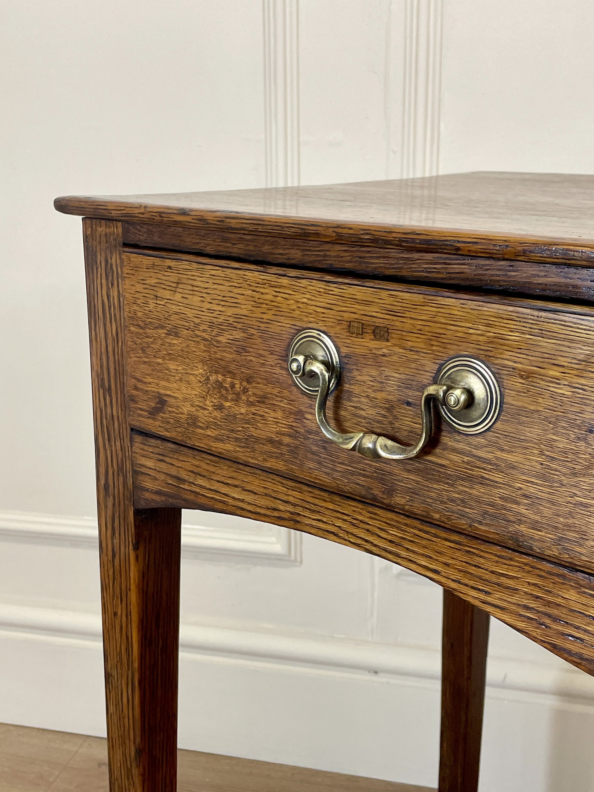 Wooden side table with a drawer and brass handle against a white paneled wall.