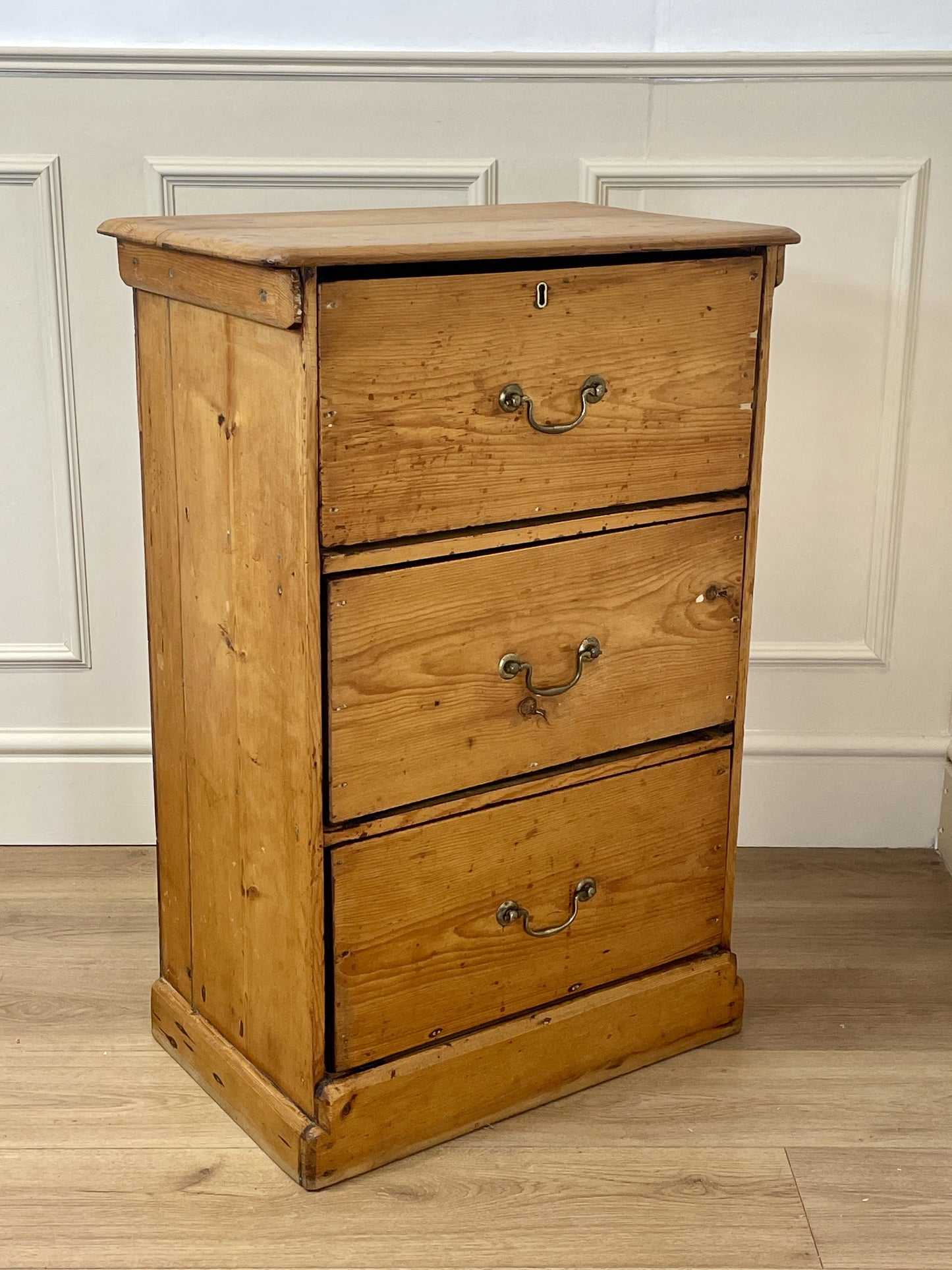 Wooden dresser with three drawers against a white paneled wall.