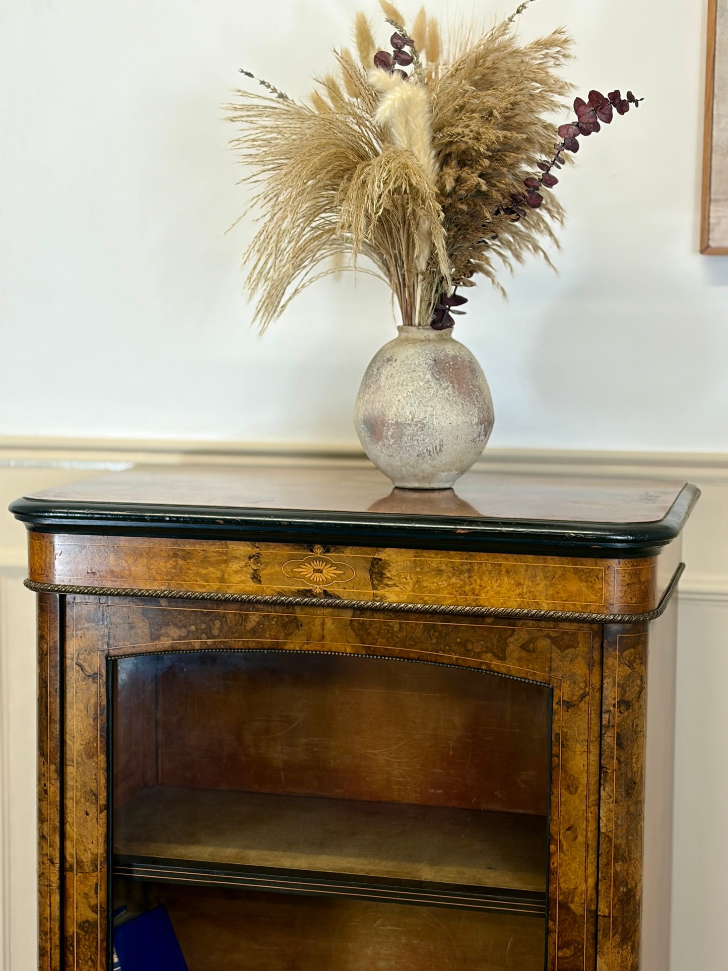 Wooden sideboard with a vase of dried plants on top against a white wall.
