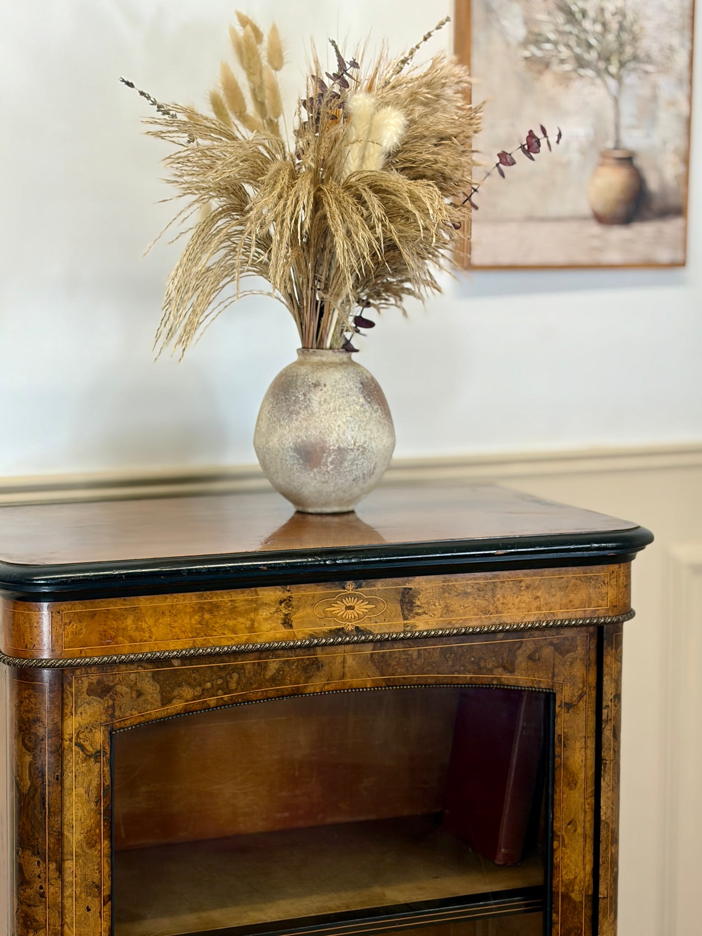 Decorative vase with dried plants on a wooden side table against a wall with a framed picture.