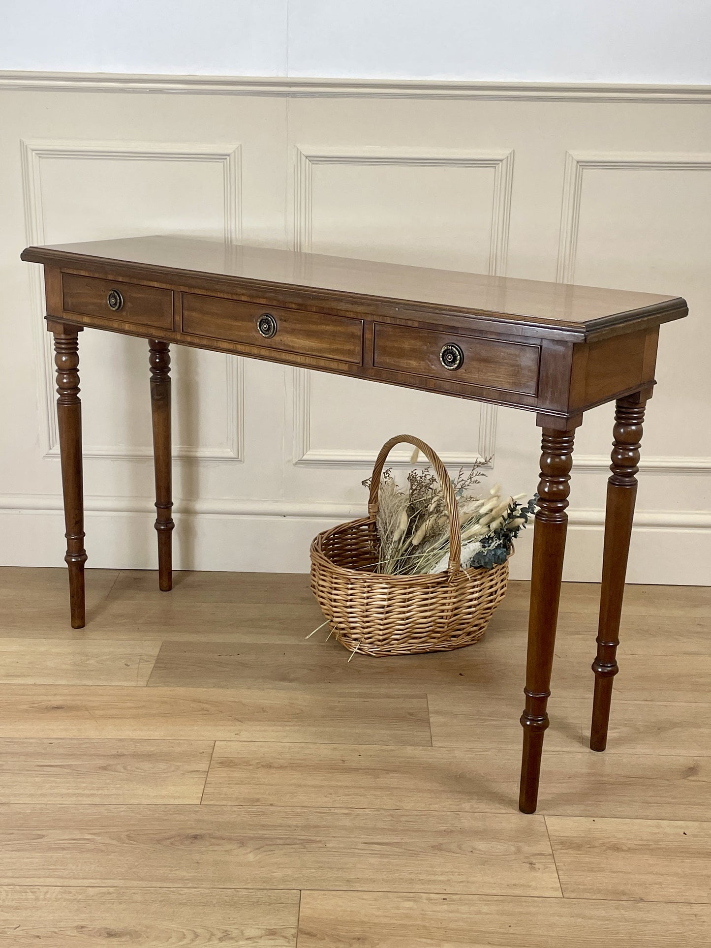 Wooden console table with a basket on a wooden floor against a white paneled wall.