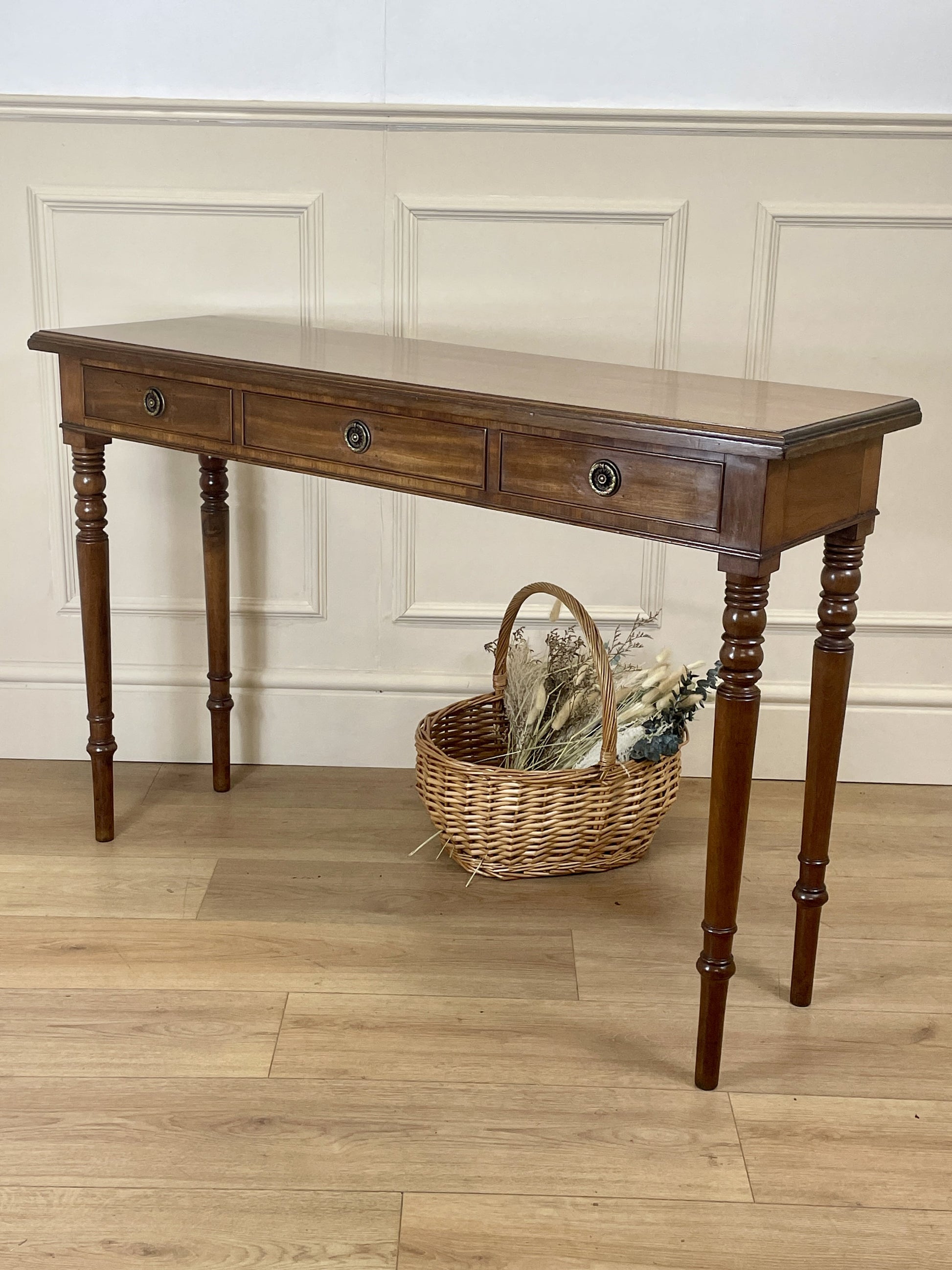 Wooden console table with a basket on a wooden floor against a white paneled wall.