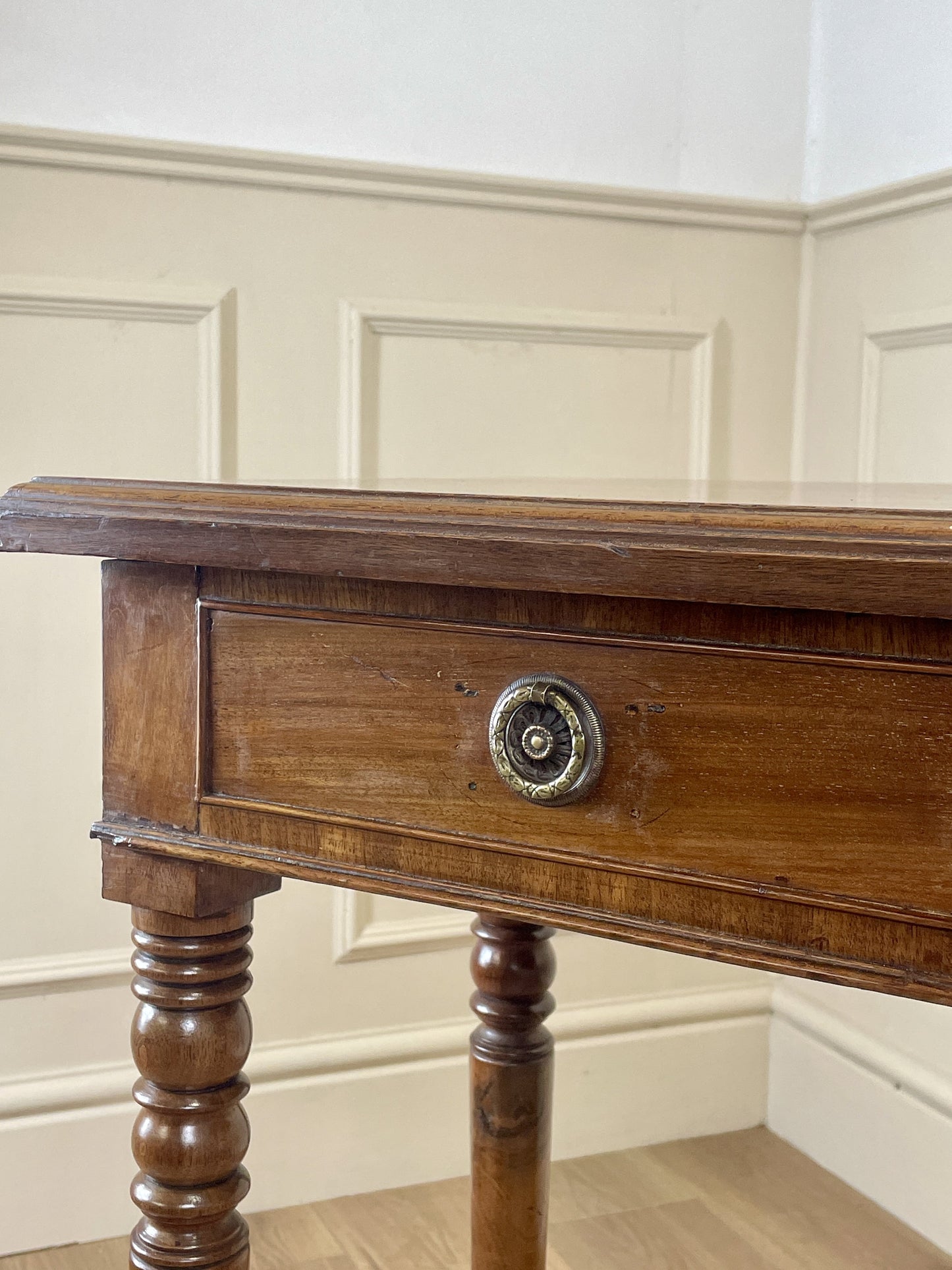 Wooden side table with a drawer and decorative knob against a white paneled wall.