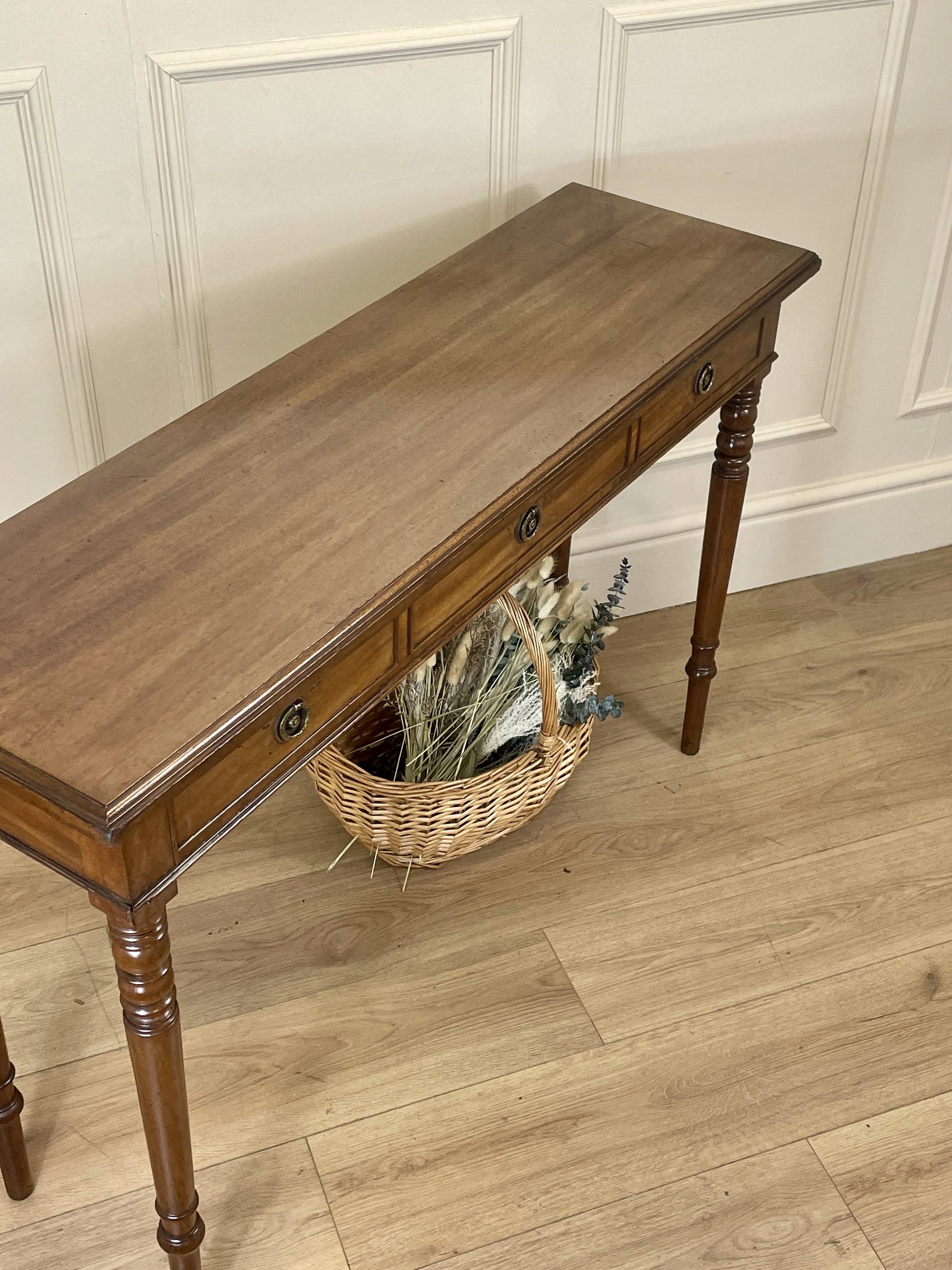 Wooden console table with a basket of flowers on a wooden floor against a paneled wall.