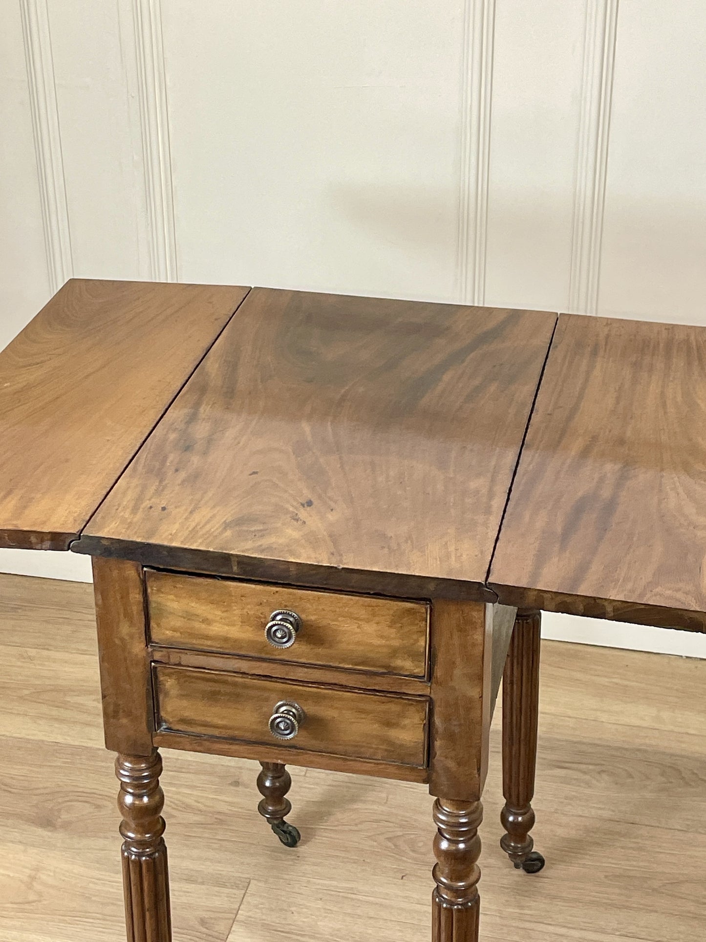 Wooden table with two drawers and a drop-leaf extension against a white paneled wall.