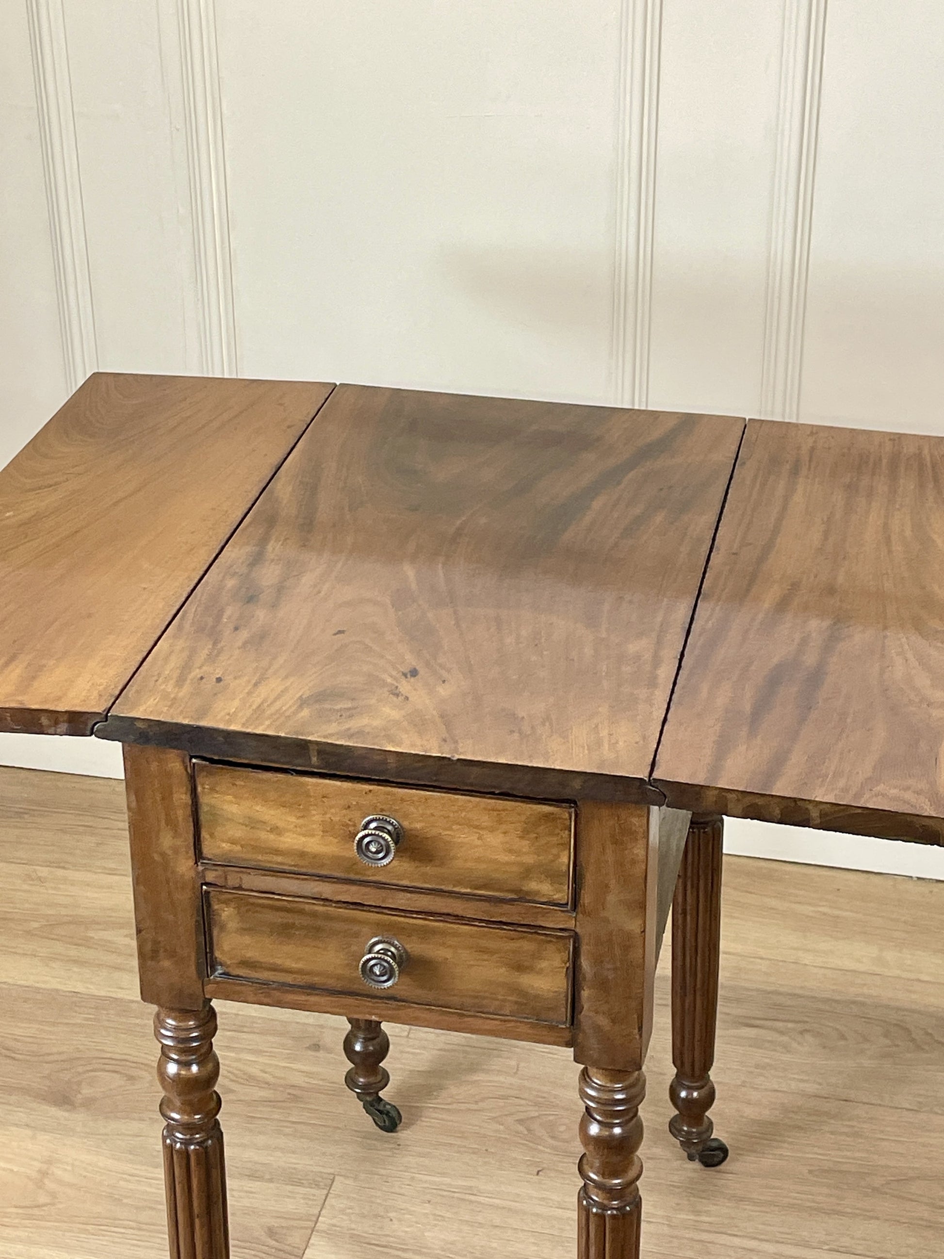 Wooden table with two drawers and a drop-leaf extension against a white paneled wall.