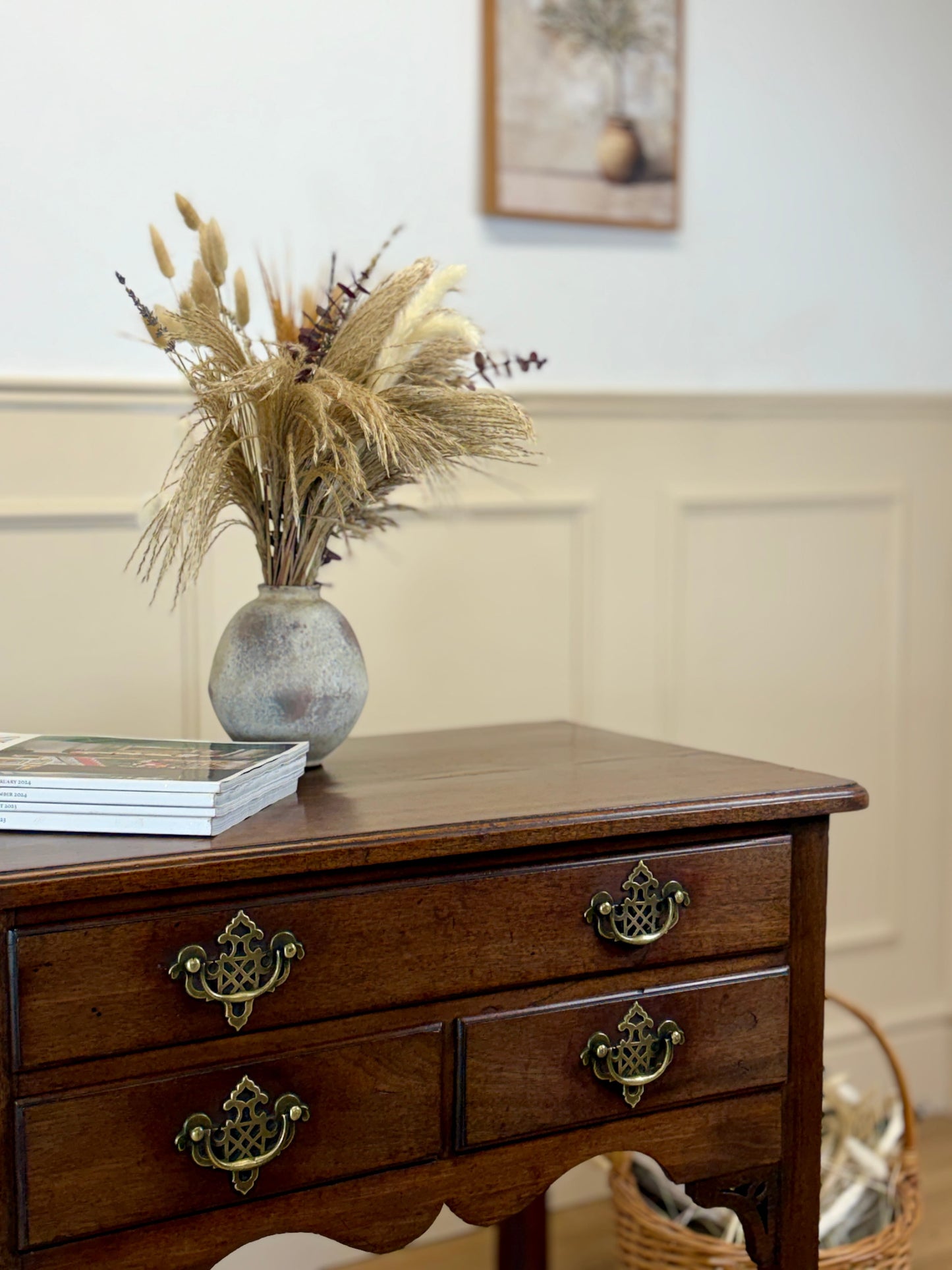 Wooden dresser with a vase of dried plants on top, against a neutral wall.
