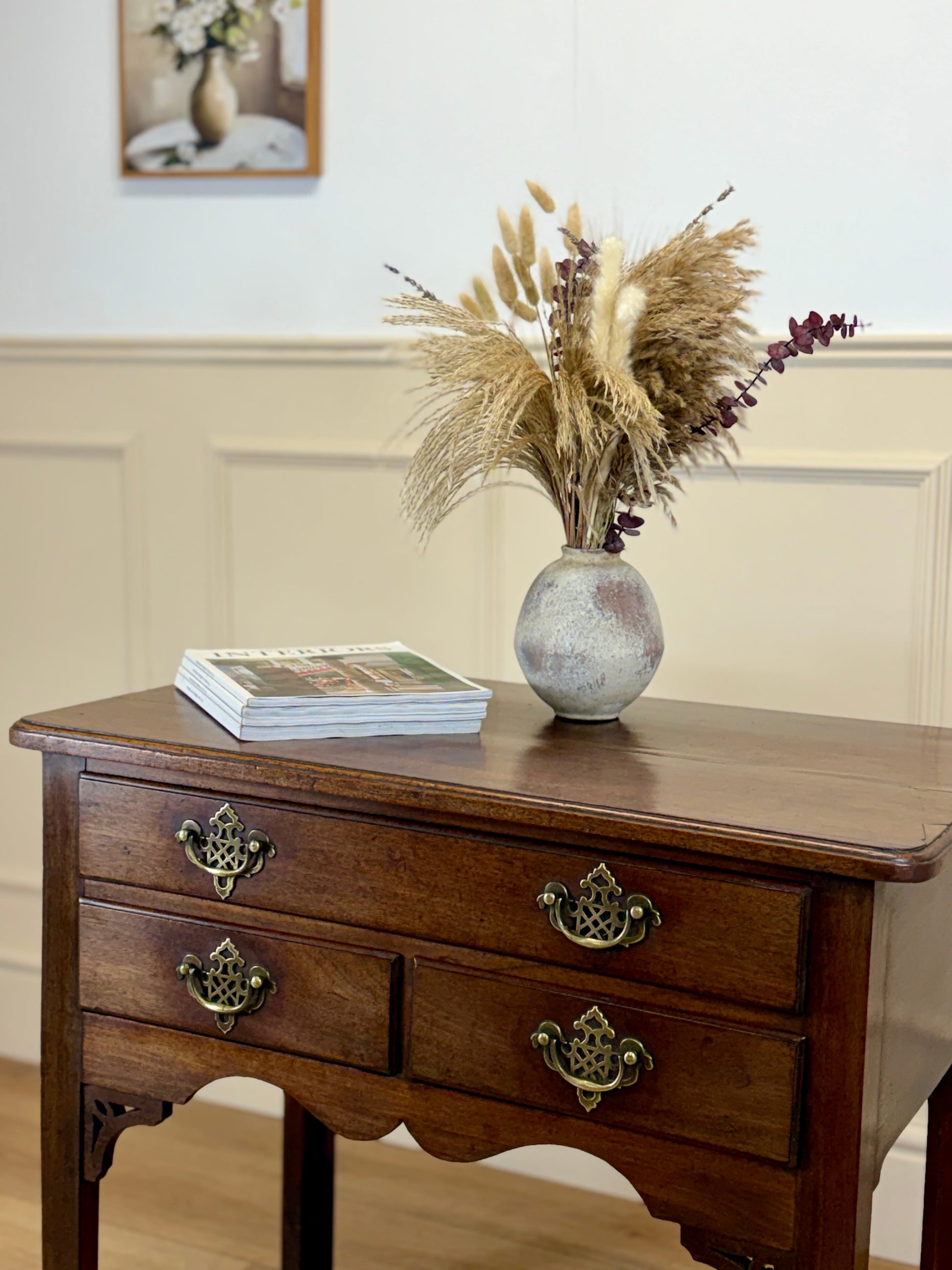 Wooden side table with a vase of dried plants and a book on a light-colored wall background