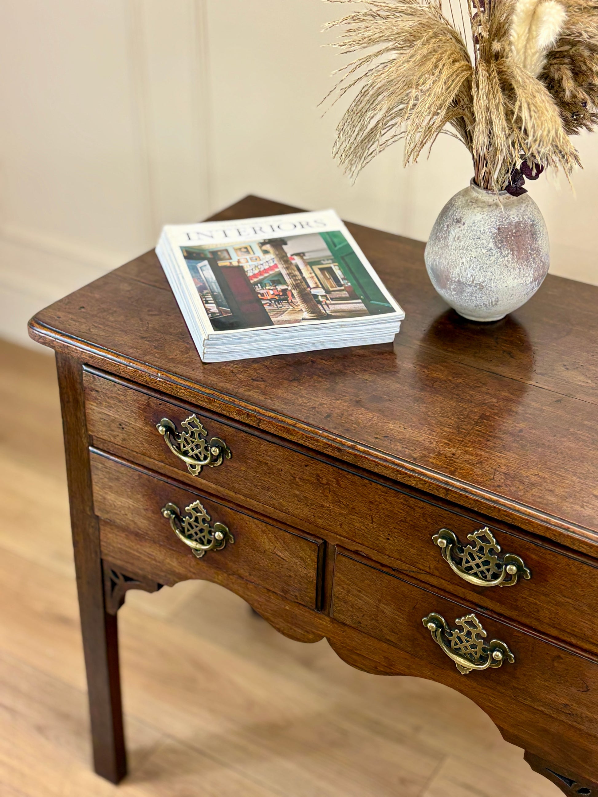 Wooden side table with two drawers, a vase with dried plants, and a magazine on a light wooden floor.