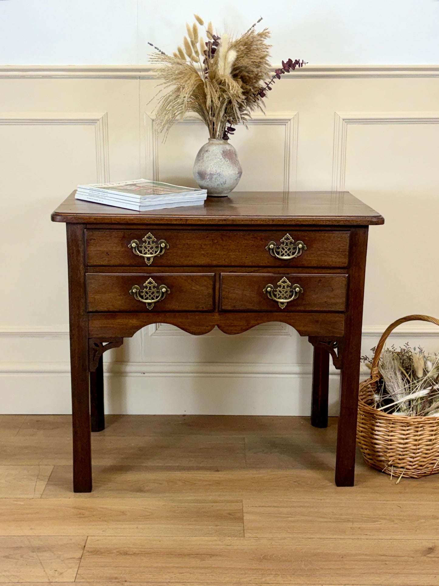 Wooden side table with two drawers, a vase of dried flowers, and a basket on a wooden floor.